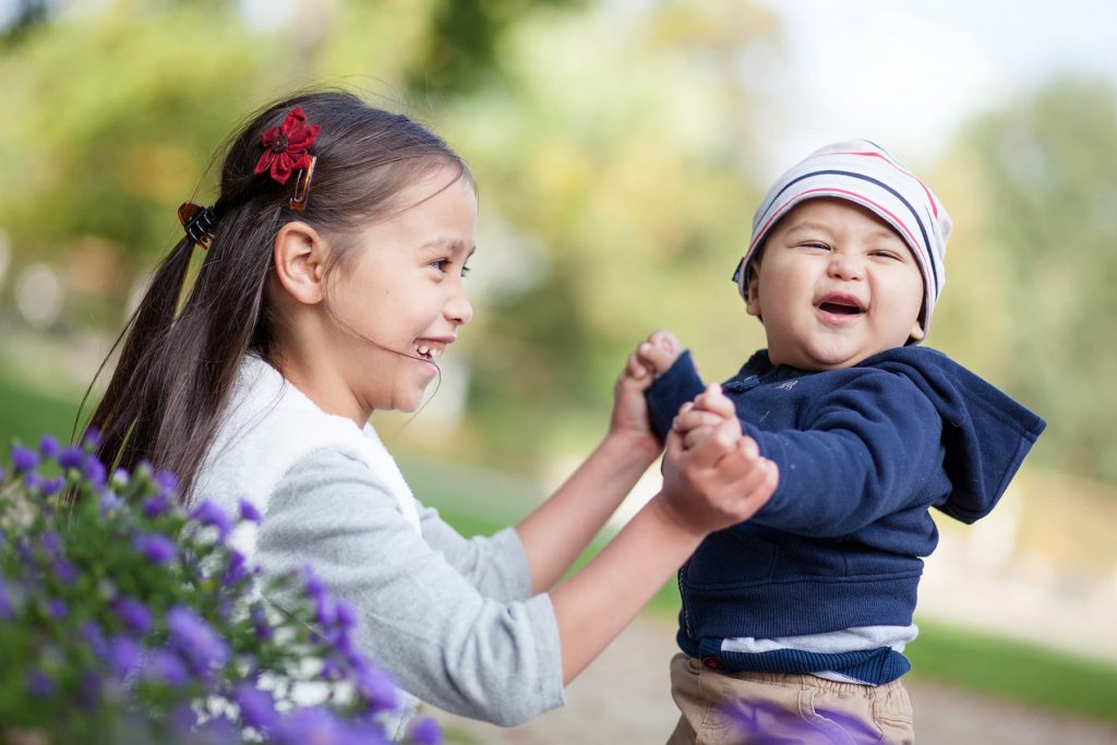 sister holds her baby brother on her lap while they laugh in the flowers