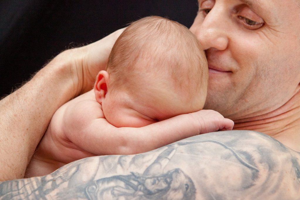 man with tattoos holds small baby daughter on his shoulder and smiles